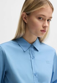 Camicia azzurra chiara con colletto, realizzata in tessuto liscio e dotata di chiusura a bottoni, con il logo ricamato "HUGO" sul lato sinistro.