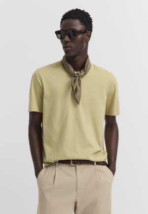 Mann trägt beiges T-Shirt, braune Hose mit dunklem geflochtenem Gürtel, gestreiftes Halstuch und Schildpatt-Sonnenbrille, steht mit den Händen in den Taschen.