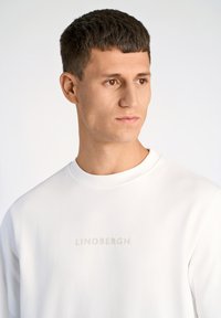 Vit sweatshirt med rund halsringning och präglad "LINDBERGH" text på bröstet, tillverkad av mjukt tyg med en slät yta.
