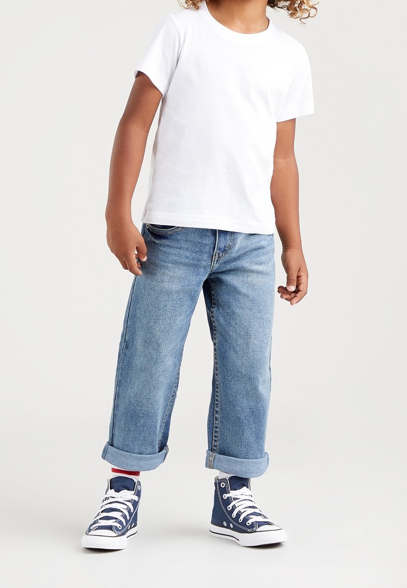 T-shirt bianco a maniche corte, jeans blu con risvolto, e sneakers alte navy con lacci bianchi. Abbigliamento casual e dalla vestibilità morbida.