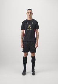 Black football jersey with gold logo and red accents, paired with black shorts and knee-length socks. Features branded footwear.
