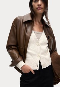 Veste en cuir marron avec un col structuré, cardigan blanc texturé avec deux boutons et pantalon noir. Texture douce et couleurs contrastées.