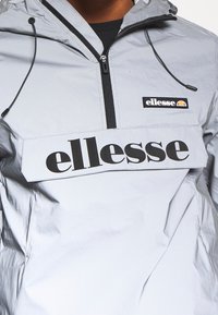Svetle šedý větrový kabát s černým páskem s logem "ellesse". Obsahuje přední zip, kapuci na stažení a hladký, lesklý povrch.