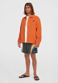 Camicia arancione con bottoni sopra una maglietta bianca, abbinata a pantaloni da nuoto neri con motivi di onde colorate e ciabatte nere.