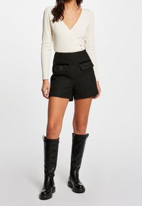 Femme portant un haut à manches longues blanc côtelé avec col en V, un short taille haute noir avec poches avant et des bottes noires montant jusqu'aux genoux.