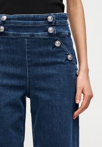 Κοντινό πλάνο σκούρου μπλε τζιν παντελονιού με πολλαπλά ασημένια διακοσμητικά κουμπιά κατά  μήκος της ζώνης και της πλευρικής ραφής, φορεμένο με μαύρο τοπ.