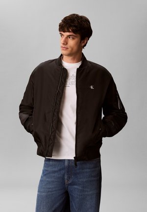 Mężczyzna noszący czarną kurtkę bomber z małym logo CK, białą koszulkę Calvin Klein, niebieskie jeansy, ręce w kieszeniach, patrzy w lewo, szare tło.