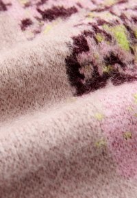 Pūkains dzijas audums maigi rozā krāsā ar purpursarkanām un dzeltenām abstraktām rakstām. Tekstūra izskatās silta un pūkaina, izceļot ērtu dizainu.