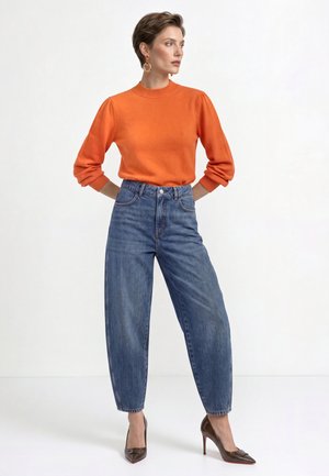 Woman standing with hands behind back wearing orange long-sleeve top, high-waisted blue jeans, gold hoop earrings, and brown high heels.