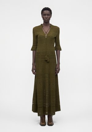 Γυναίκα ντυμένη με μάξι πλεκτό φόρεμα σε ελαιώδες πράσινο με σχέδια, μανίκια τριών τετάρτων και καφέ μποτάκια μέχρι τον αστράγαλο, στέκεται μπροστά σε λευκό φόντο.