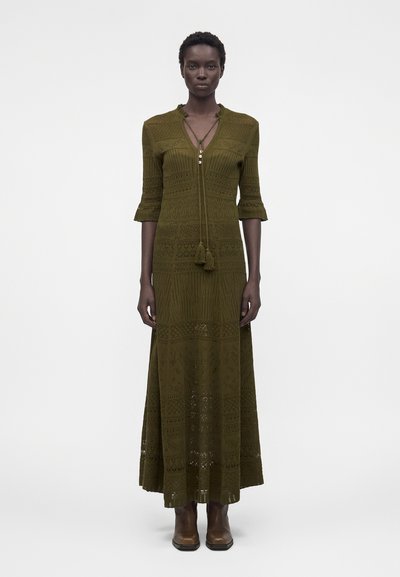 Femme portant une robe maxi en maille à motif vert olive avec des manches trois-quarts et des bottines marron, debout devant un fond blanc.