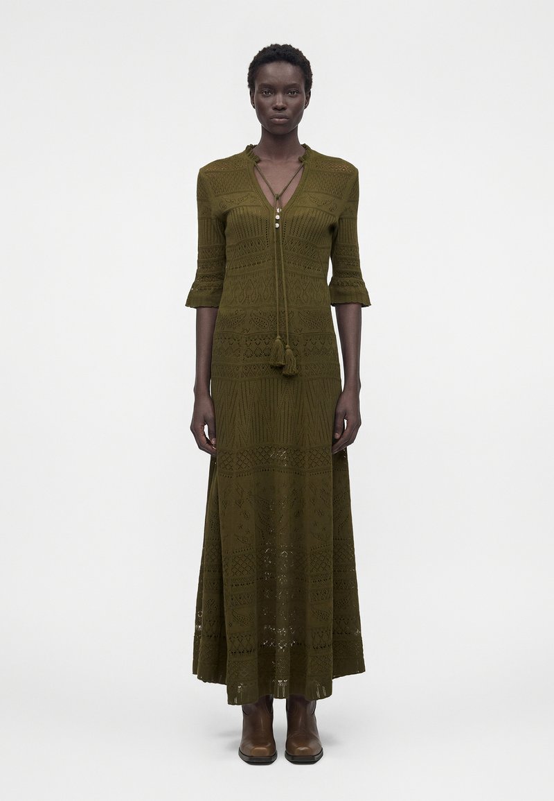 Γυναίκα ντυμένη με μάξι πλεκτό φόρεμα σε ελαιώδες πράσινο με σχέδια, μανίκια τριών τετάρτων και καφέ μποτάκια μέχρι τον αστράγαλο, στέκεται μπροστά σε λευκό φόντο.