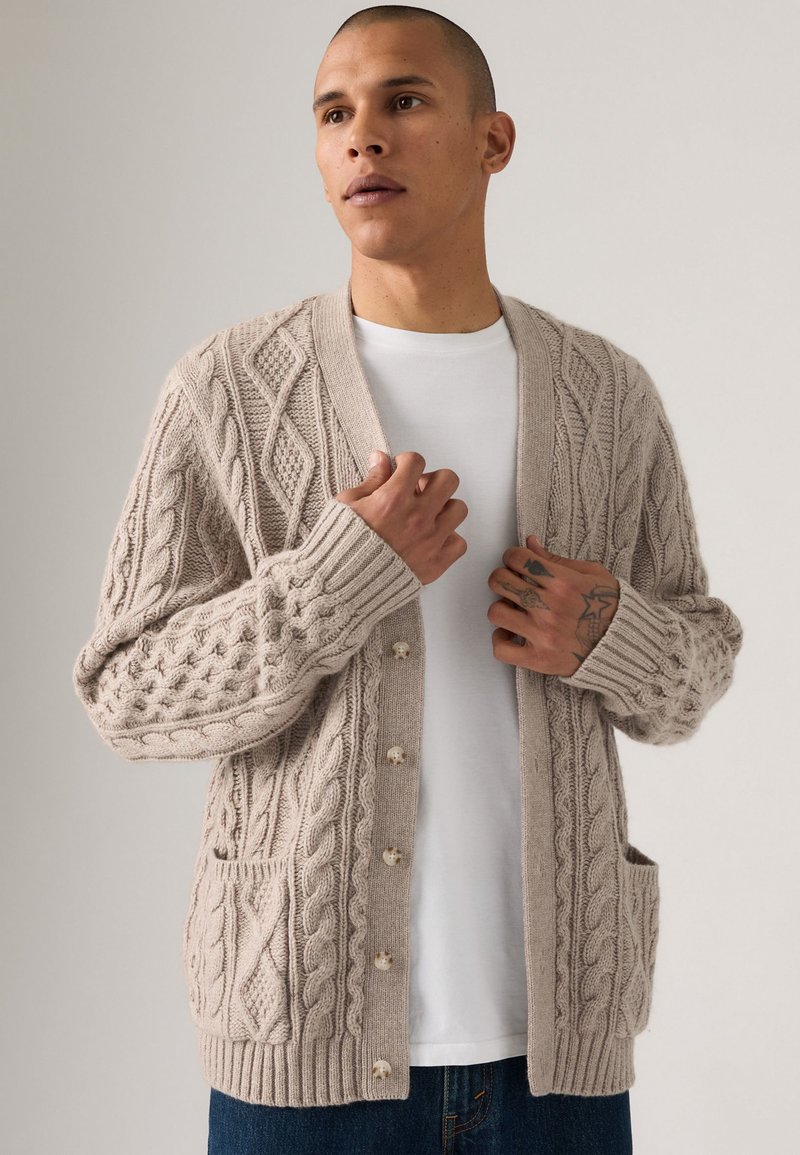 Light beige knitted cardigan with intricate cable patterns, ribbed cuffs, and three buttons. Features a front pocket. Worn over a white shirt.