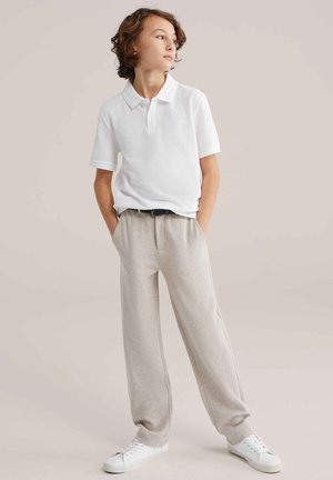 Chlapec so stredne dlhými kučeravými vlasmi oblečený v bielej polokošeli, béžových nohaviciach a bielych teniskách stojaci s rukami v vreckách.