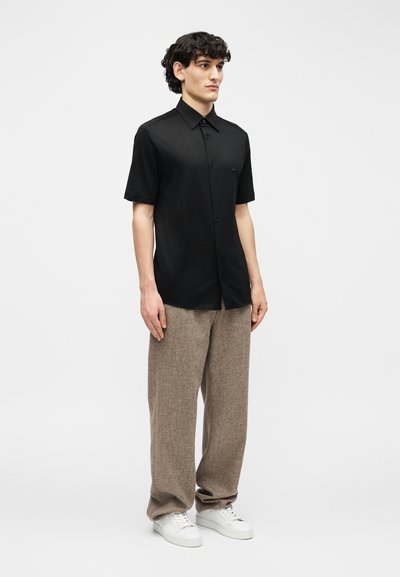 Άνδρας που φοράει μαύρο κοντομάνικο πουκάμισο με κουμπιά, ανοιχτόχρωμο μπεζ χαλαρό παντελόνι και λευκά αθλητικά παπούτσια, στέκεται μπροστά σε απλό ανοιχτό γκρι φόντο.