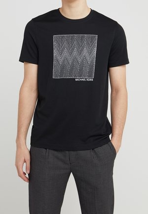 Mann trägt ein schwarzes T-Shirt mit kurzen Ärmeln, das eine weiße Zickzack-Quadrat-Grafik und den Text "MICHAEL KORS" zeigt, kombiniert mit schwarz-weiß karierten Hosen.
