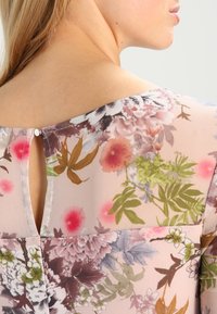 Blusa floral con una tela rosa semitransparente, que presenta una apertura en forma de llave en la parte posterior y patrones florales en tonos morado, verde y rojo.