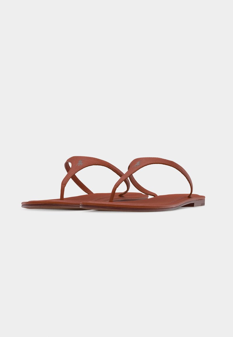 Sandales plates en cuir marron avec fines lanières sur les orteils et logo discret sur la lanière, présentées sur un fond blanc uni.