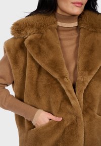 Gilet in pelliccia sintetica marrone con ampio colletto, chiusura con un bottone e tasche frontali. Indossato sopra un maglione a collo alto marrone chiaro.