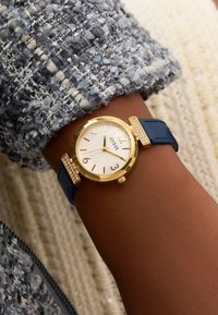Montre en or avec cadran blanc, bracelet en cuir bleu et accents en cristal sur les cornes. Positionnée sur un poignet contre un tissu texturé.