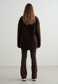 Bruine, oversized fleece trui met een gestructureerd oppervlak, gecombineerd met strakke, bruine broek en witte sneakers. Zicht vanuit de rug.