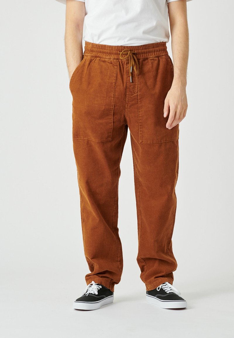 Homme portant un pantalon en velours côtelé couleur rouille avec taille élastique et cordon, baskets noires avec lacets blancs et un t-shirt blanc, debout.