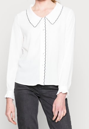 Kvinde iført en hvid bluse med lange ærmer og bølget sort kant på krave, knaplinje og manchetter, kombineret med mørkegrå jeans.