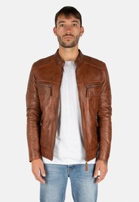 Giacca di pelle marrone con colletto rialzato, tasche con zip e finitura testurizzata, abbinata a una t-shirt bianca e jeans blu.