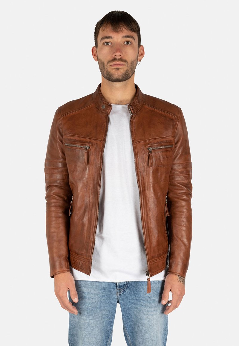 Giacca di pelle marrone con colletto rialzato, tasche con zip e finitura testurizzata, abbinata a una t-shirt bianca e jeans blu.