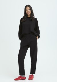 Melna kapuce un saskaņotas bikses, abas izgatavotas no mīkstas auduma. Modelis valkā sarkanus sporta apavus ar pelēkām zeķēm, demonstrējot brīvu piegriezumu un vienkāršu dizainu.