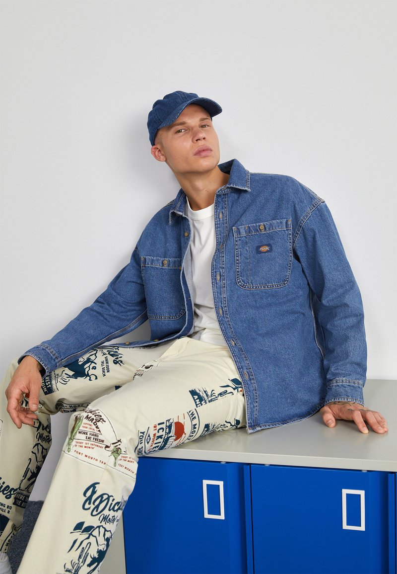 Μπλε τζιν πουκάμισο με δύο τσέπες στο στήθος, συνδυασμένο με κρεμ παντελόνι που έχει διάφορες μπλε και μ�αύρες επιγραφές και γραφικά. 