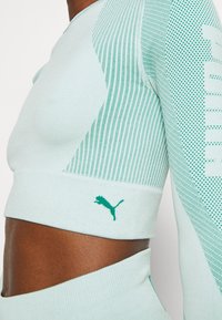 Hellblau-grünes Sportoberteil mit strukturierten Netzbereichen, kontrastierendem grünen Logo und gerippten Manschetten. Enganliegendes Design mit cropped Stil.