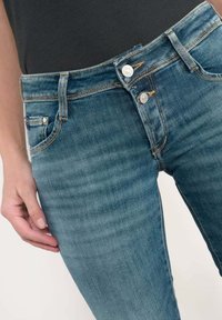 Jeans en denim bleu clair, avec une coupe slim, deux poches frontales, deux fermetures à bouton et des détails de couture visibles. Texture douce.