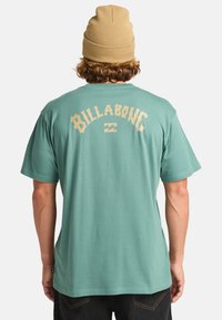 Camiseta de manga corta de color verde menta con el logotipo "Billabong" en color beige impreso en la parte posterior. Tejido suave con un ajuste relajado, diseño simple y cuello redondo.