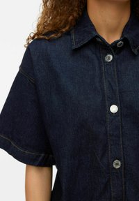 Mörk denimskjorta med korta ärmar, med en knappstängning framtill, kontrasterande gul sömnad och metalliska tryckknappar.