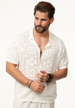 Weißes Kurzarmhemd mit einem filigranen offenen Strickmuster, das einen Kragen und eine Knopfleiste aufweist. Begleitende dekorative Armbänder sind sichtbar.