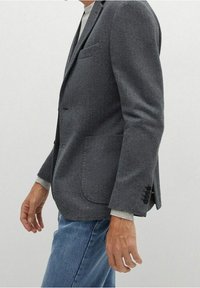 Uomo che indossa un blazer grigio a spina di pesce sopra un maglione grigio chiaro e jeans blu, in piedi contro uno sfondo chiaro e semplice.