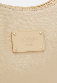 Sac beige texturé avec une étiquette beige rectangulaire attachée par quatre rivets métalliques, gravée du texte "JOOP! JEANS".