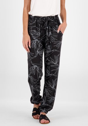 Pantalons noirs avec impression botanique blanche, taille élastique avec cordon de serrage, poches latérales et poignets élastiques aux chevilles. Portés avec des sandales noires.