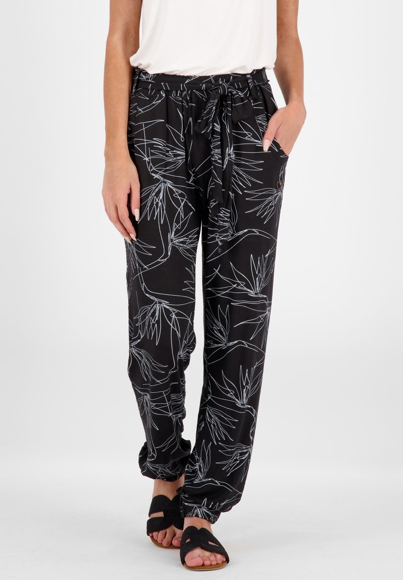 Schwarze Hose mit weißem botanischem Druck, elastischem Bund mit Kordelzug, Seitentaschen und elastischen Säumen an den Knöcheln. Getragen mit schwarzen Sandalen.