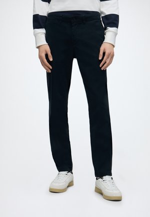 Άνδρας που φοράει μαύρο παντελόνι slim fit, λευκά αθλητικά παπούτσια και λευκό πουκάμισο με μακριά μανίκια και μαύρες οριζόντιες ρίγες.
