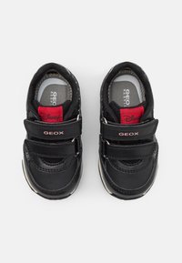 Svarta läderskor för barn med kardborreband, gummisula och en röd och vit logotyp på hälen. Slät textur och rund form.