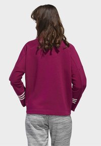 Person med krulligt brunt hår som bär en magenta sweatshirt med vita ränder på ärmarna och gråmelerade byxor, vänd bort från kameran.