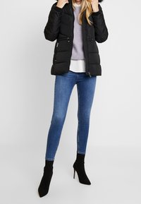 Chaqueta acolchada negra con capucha, cremalleras y paneles contrastantes, llevada sobre un suéter lavanda y una blusa blanca, combinada con vaqueros azules y botas negras de tobillo.