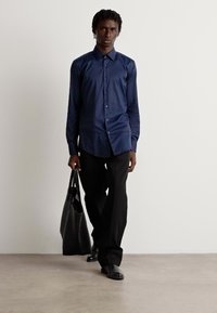 Camisa de manga larga azul marino con cierre de botones, combinada con pantalones y zapatos negros. El modelo lleva una bolsa tote negra y está de pie frente a un fondo neutro.