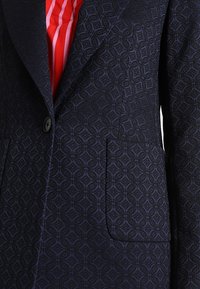 Granatowa marynarka z teksturowanym geometrycznym wzorem, z wyłogiem z wcięciem, zapinana na guziki, z dwoma przednimi kieszeniami. Czerwono-biały paskowany podszewka.