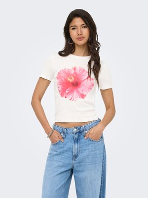 Mujer con camisa blanca de manga corta con gran estampado de flor rosa, vaqueros azules de talle alto y pendientes de aro coloridos, manos en los bolsillos.