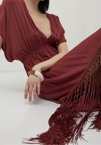 Maroon dress with a deep V-neck and gathered waist, featuring a textured fabric and fringed hem. A chunky, two-tone bracelet adorns the wrist.