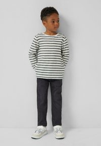 Een jong kind staat tegen een effen muur en draagt een wit met zwart gestreept shirt met lange mouwen, donkere broek en witte sneakers.