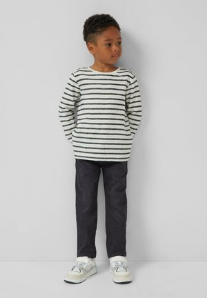 Een jong kind staat tegen een effen muur en draagt een wit met zwart gestreept shirt met lange mouwen, donkere broek en witte sneakers.